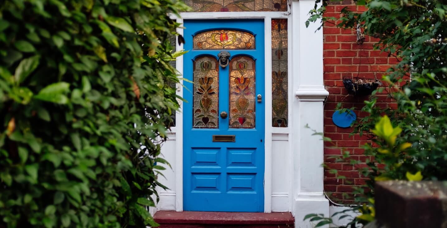 Blue door of a small office shop with stained glass windows tucked away behind a lush green plants.